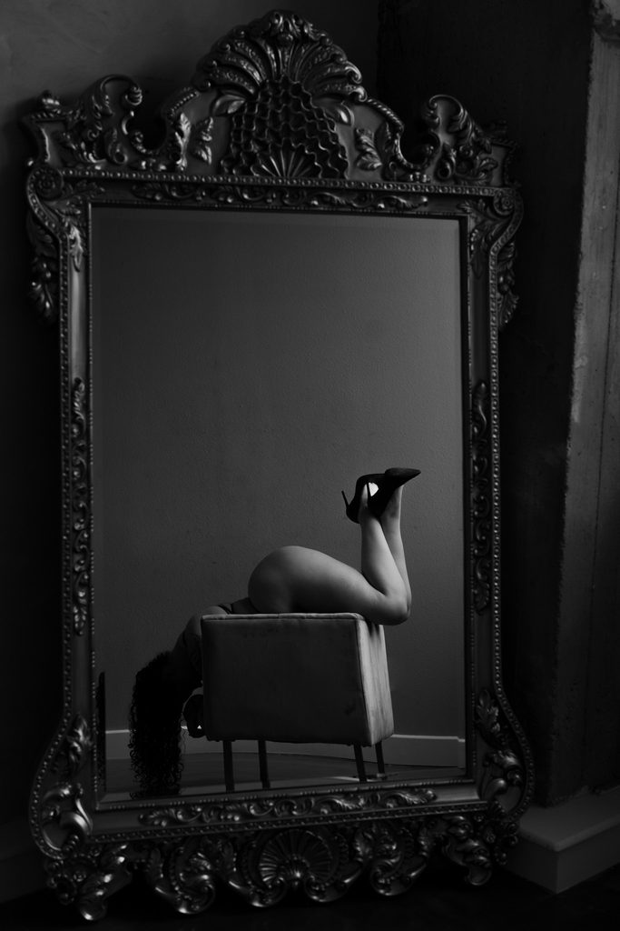 Black-and-white boudoir pose reflected in an ornate mirror, featuring a woman reclining over a chair with her legs raised in high heels.